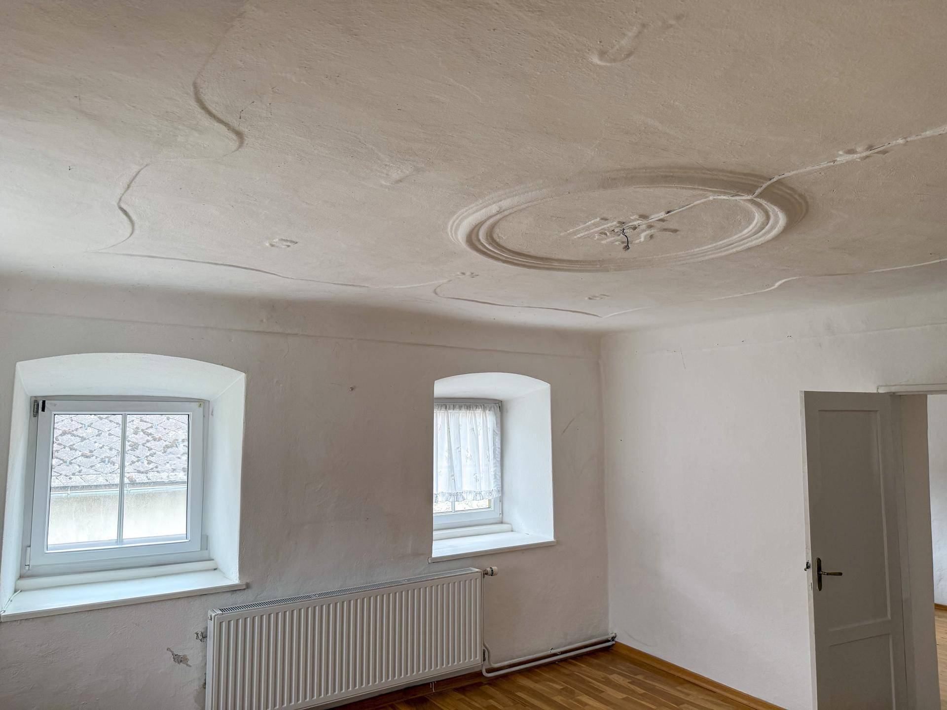 Helles Zimmer mit Holzboden, Stuckdecke und zwei Fenstern, bereit zur individuellen Gestaltung.