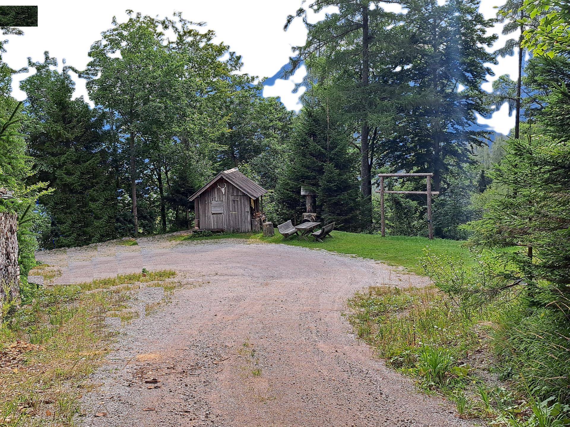 Eine kleine Holzhütte in einer Lichtung mit einem Schotterplatz und umgebenden Bäumen, inklusive Sitzgelegenheit.
