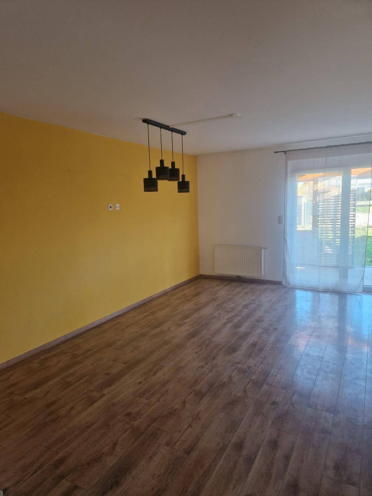 Heller Wohnraum mit Laminatboden, gelber Akzentwand und Zugang zur Terrasse.