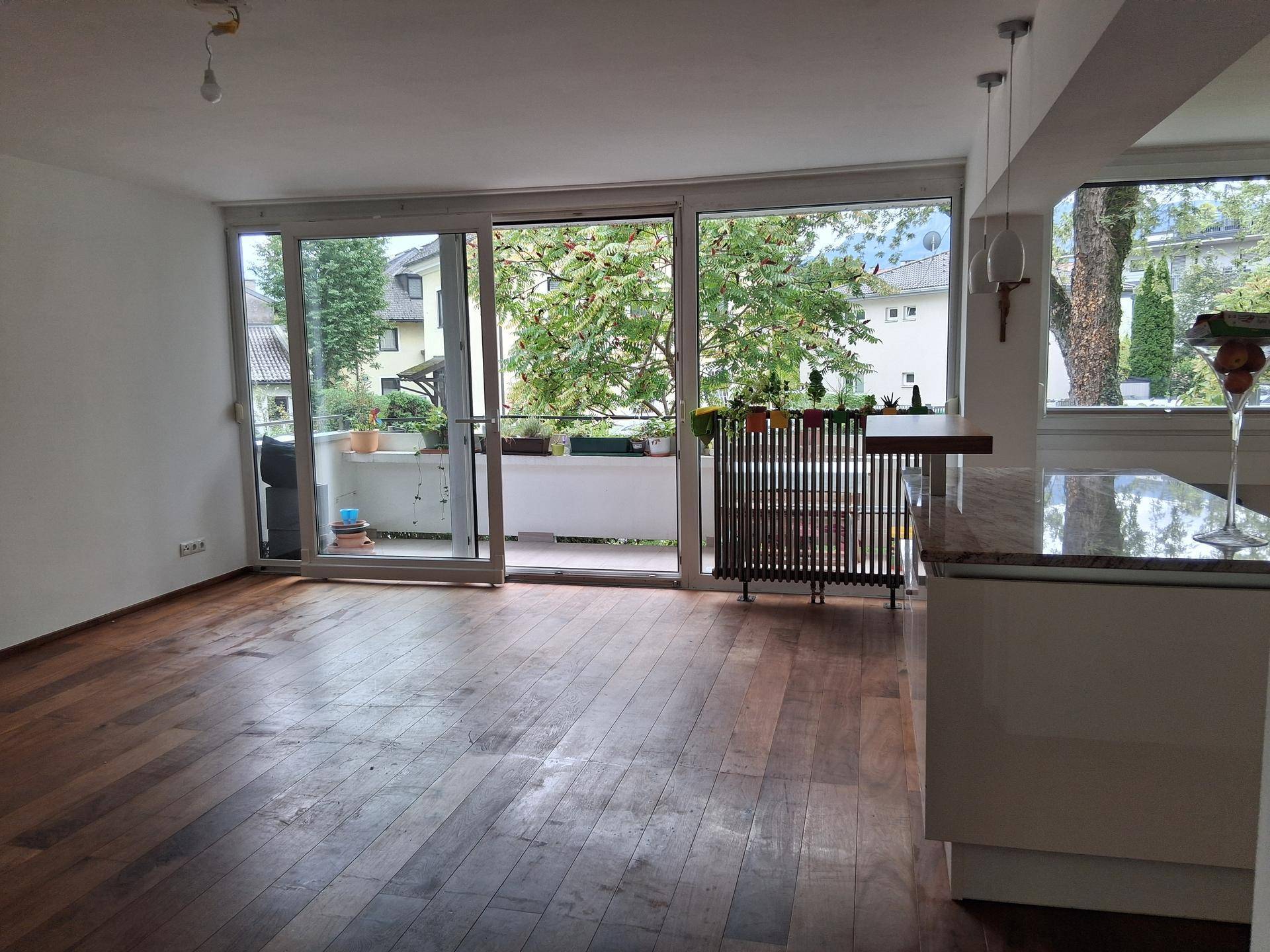 Heller offener Wohnbereich mit Holzboden und großem Schiebefenster zum Balkon mit Blick ins Grüne.