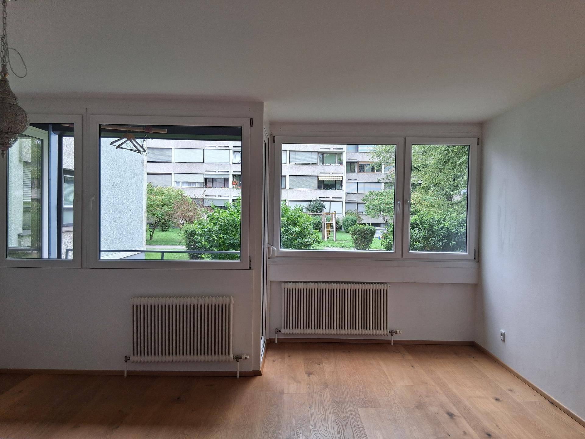 Geräumiges Zimmer mit Holzboden, großen Fenstern und einem angenehmen Blick ins Grüne.