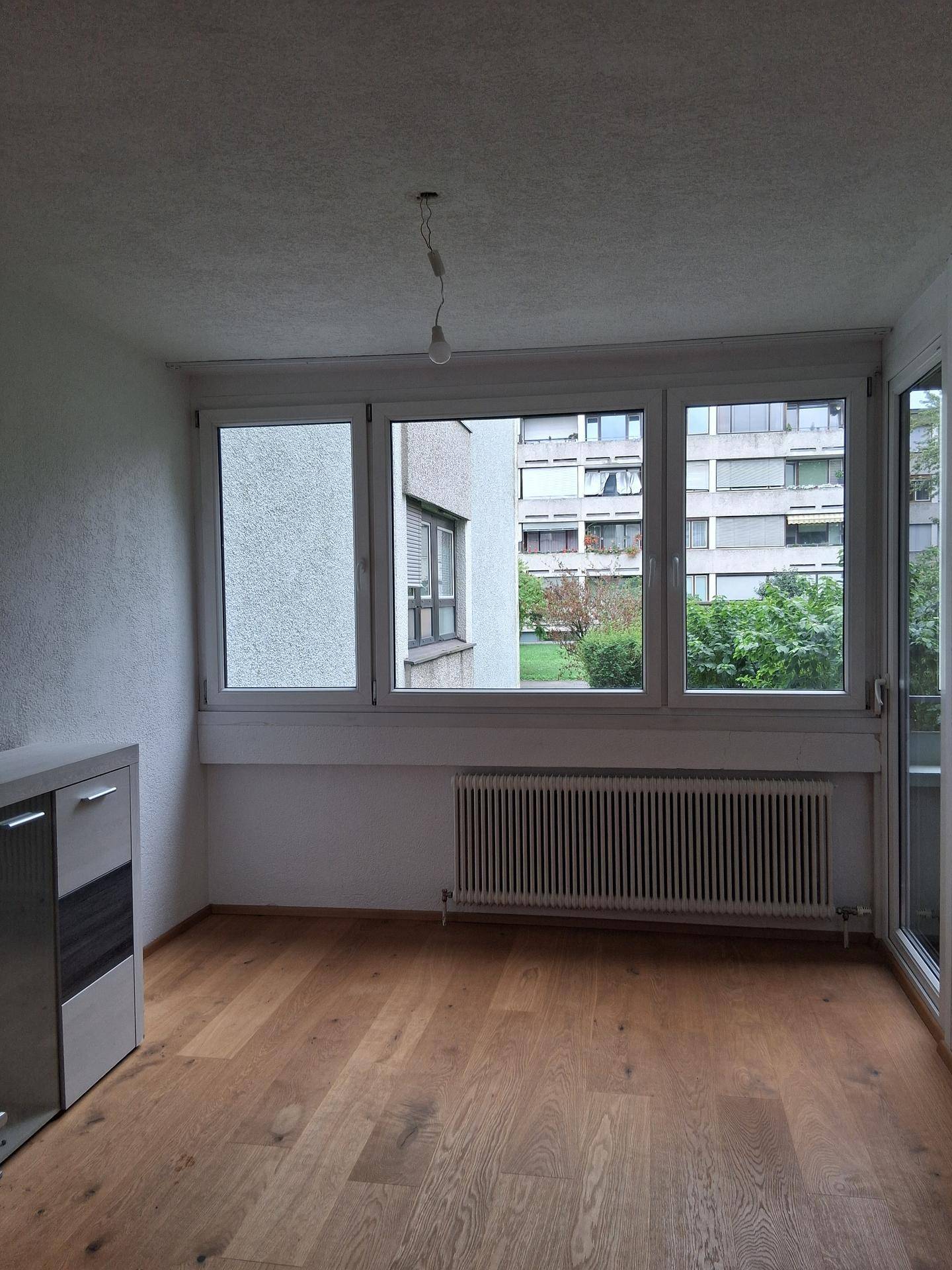Helles Zimmer mit Parkettboden, Heizkörper und Blick auf die umliegenden Gebäude und Grünflächen.