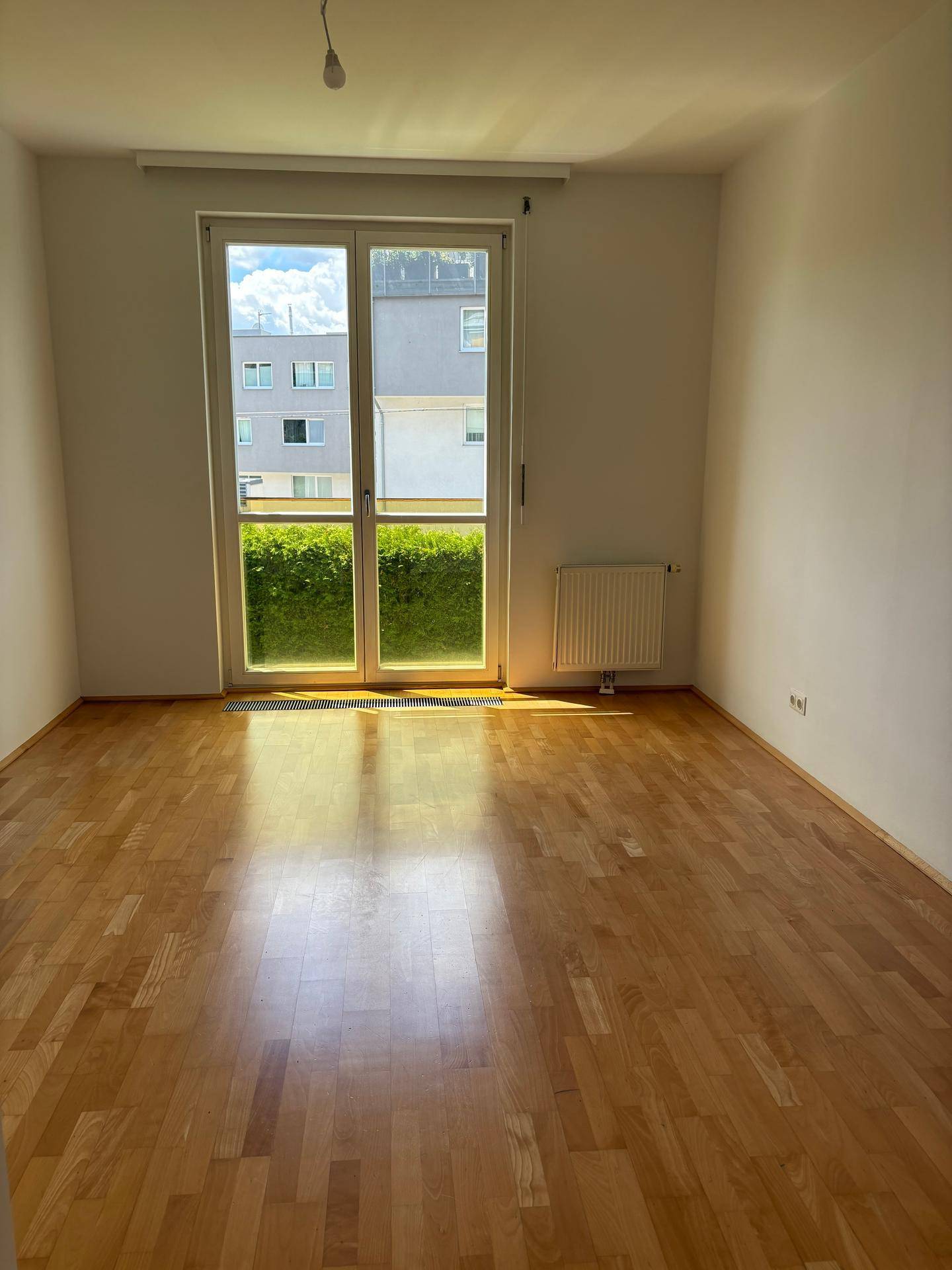 Heller Raum mit Holzparkett, großem Fenster und Blick ins Grüne, ausgestattet mit Heizkörper.