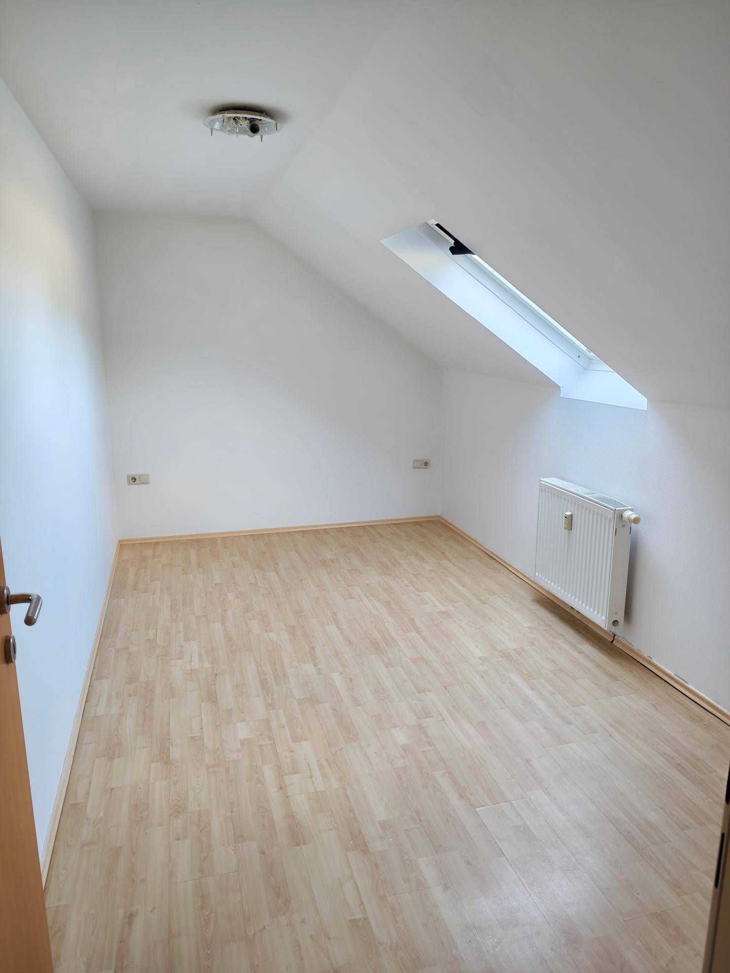 Helles Zimmer mit Dachschräge, Laminatboden, Heizkörper und einem Dachfenster für natürliches Licht.