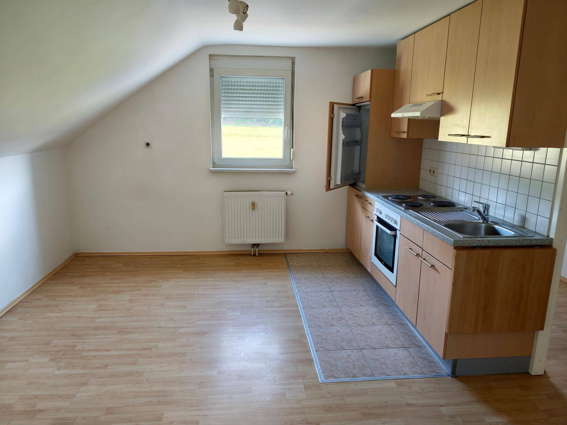 Offener Wohnbereich mit kleiner Küchenzeile, Fenster und Heizkörper unter einer Dachschräge.