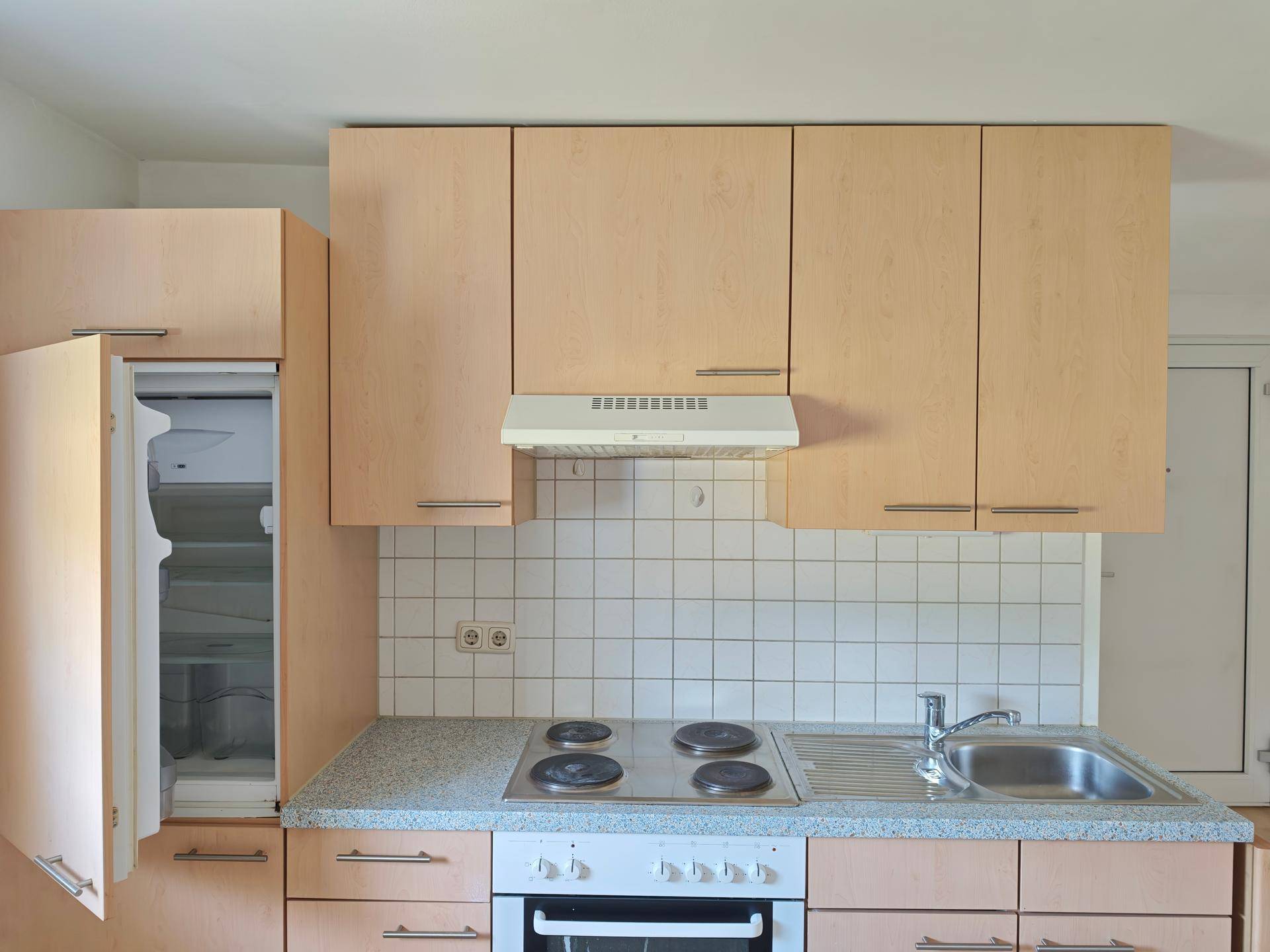 Einfache Einbauküche mit hellen Holzfronten, Elektroherd, Spüle und integriertem Kühlschrank.