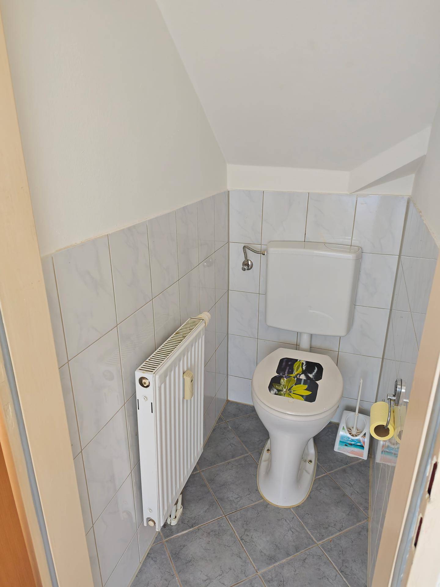 Kleines Gäste-WC mit Toilette, Heizkörper und hellgrauen Bodenfliesen unter einer Dachschräge.
