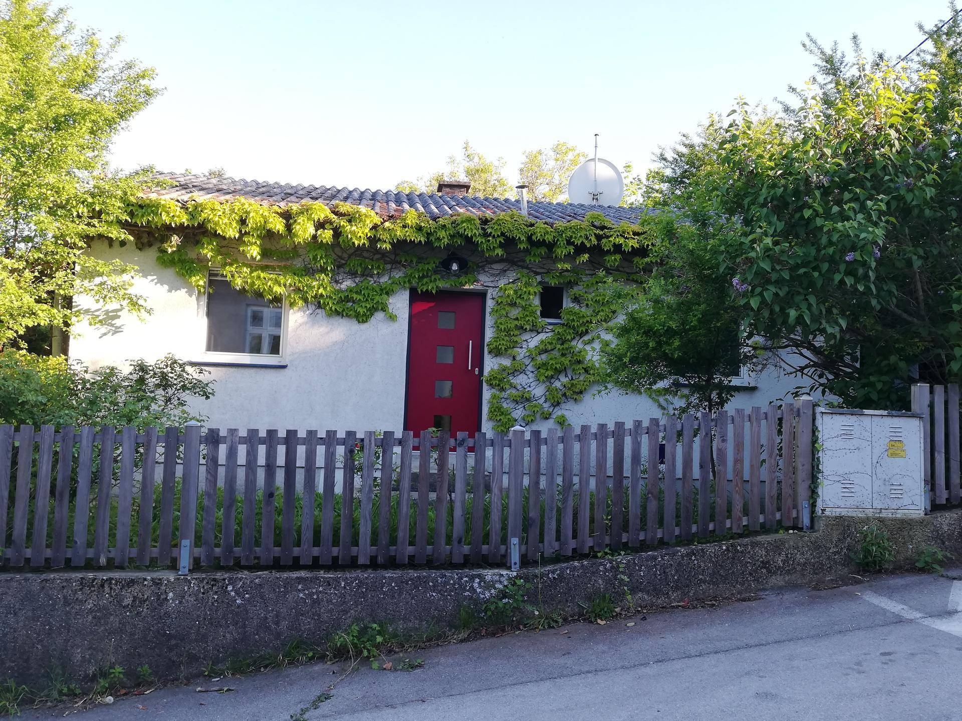 Vorderansicht des Hauses mit roter Eingangstür, Holzzaun und bewachsenen Fassadenbereichen.