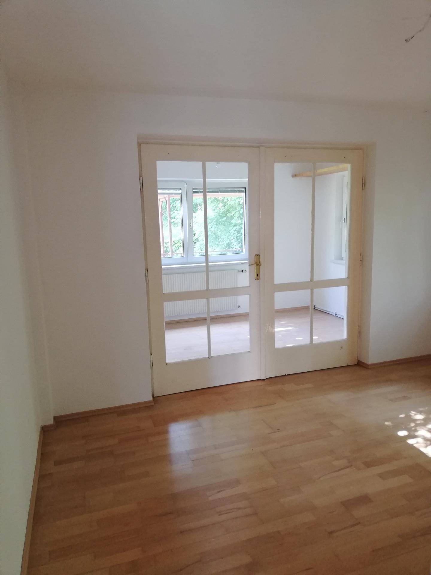 Heller Raum mit Holzboden und Doppeltüren, die zu einem weiteren Zimmer führen.