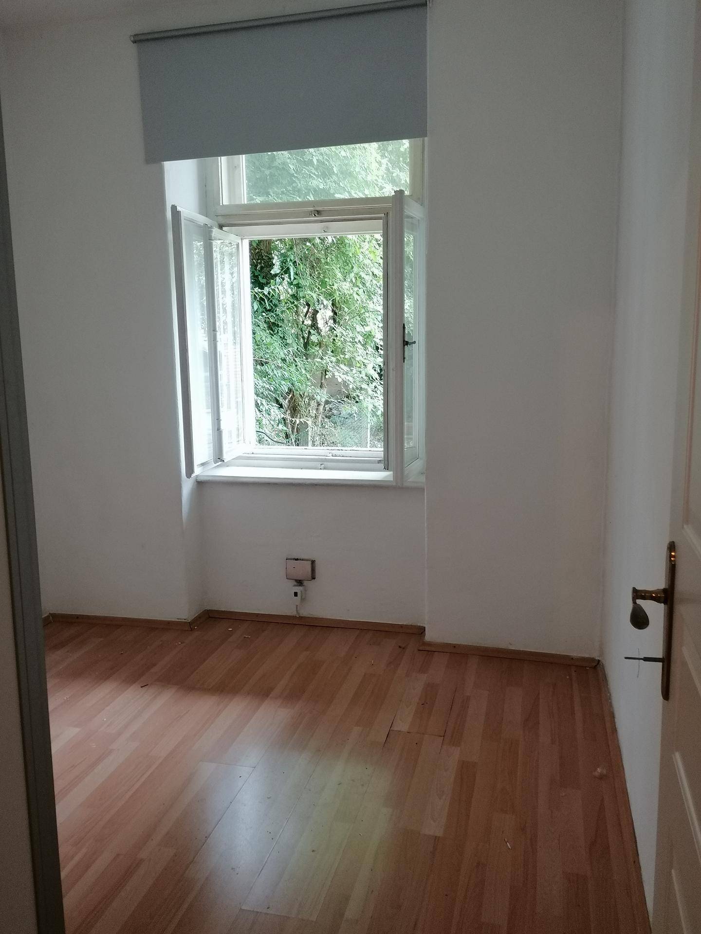 Ruhiges Zimmer mit Holzboden und Blick ins Grüne durch das geöffnete Fenster.