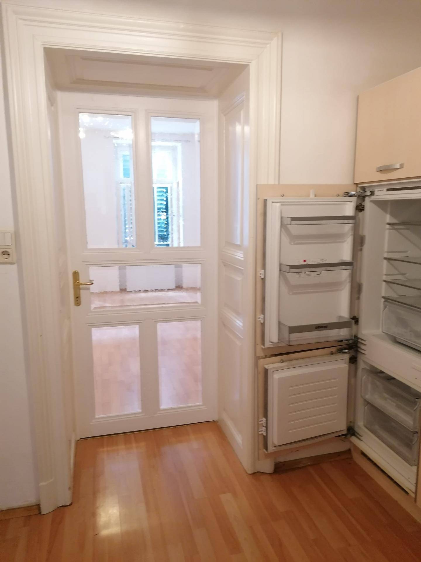 Blick aus der Küche mit offenem Kühlschrank durch eine Glastür in einen hellen Raum.