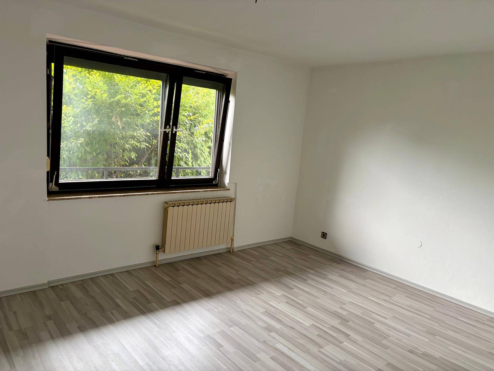 Helles Zimmer mit Laminatboden, Heizkörper und Fenster mit Blick ins Grüne.