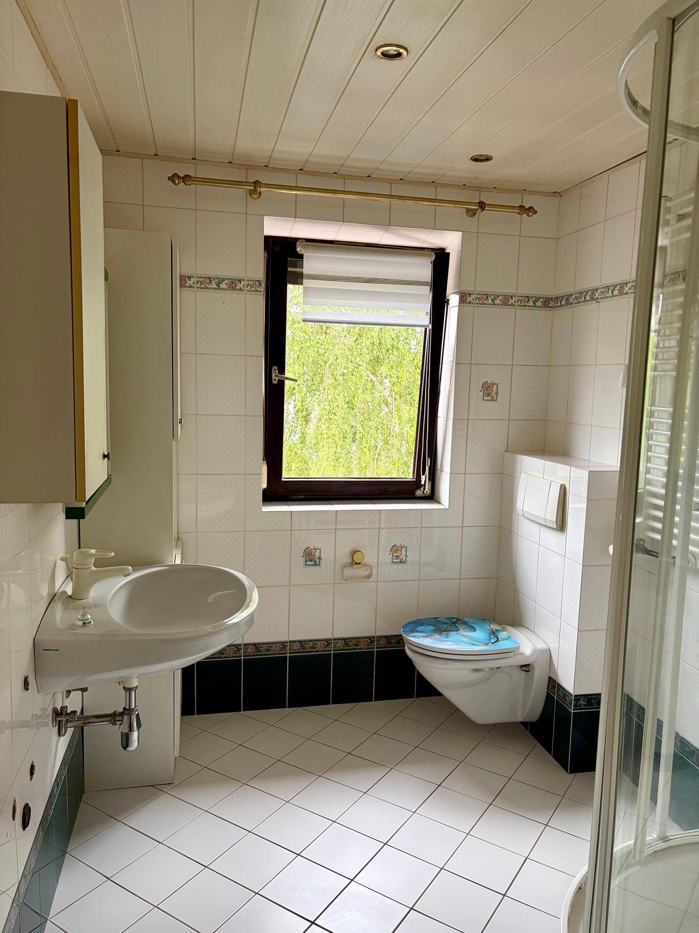 Gefliestes Badezimmer mit Fenster, Dusche, WC und Waschbecken in hellem Design.