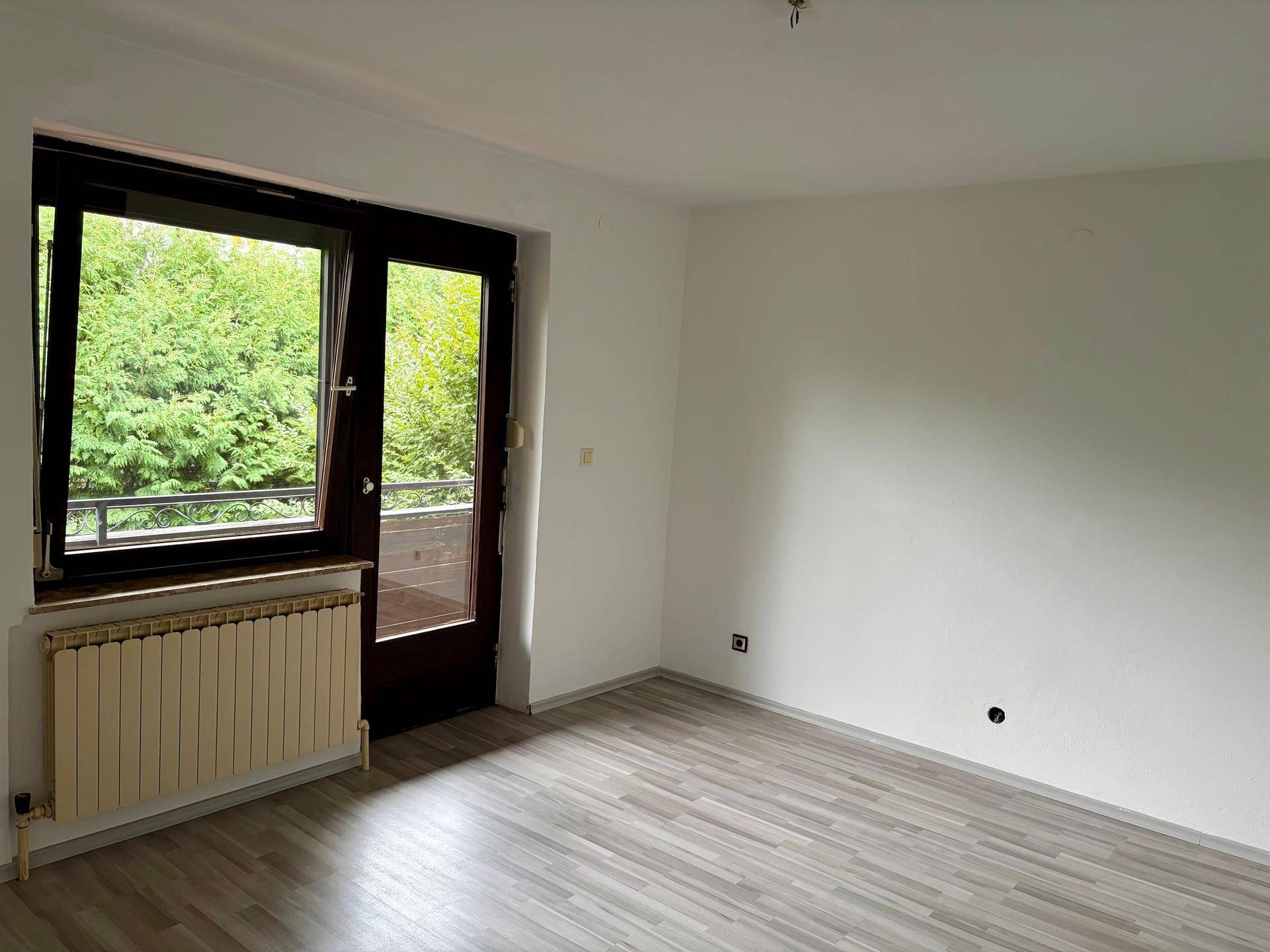 Helles Zimmer mit Laminatboden, Heizkörper und Zugang zu einem Balkon mit Grünblick.