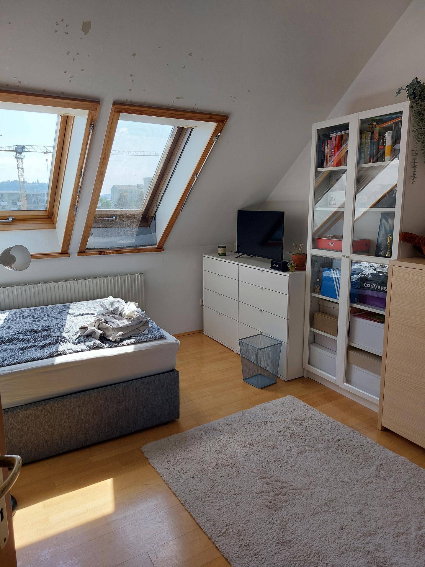 Helles Schlafzimmer mit Bett, Kommode, Bücherregal und großen Dachfenstern in der Dachschräge.
