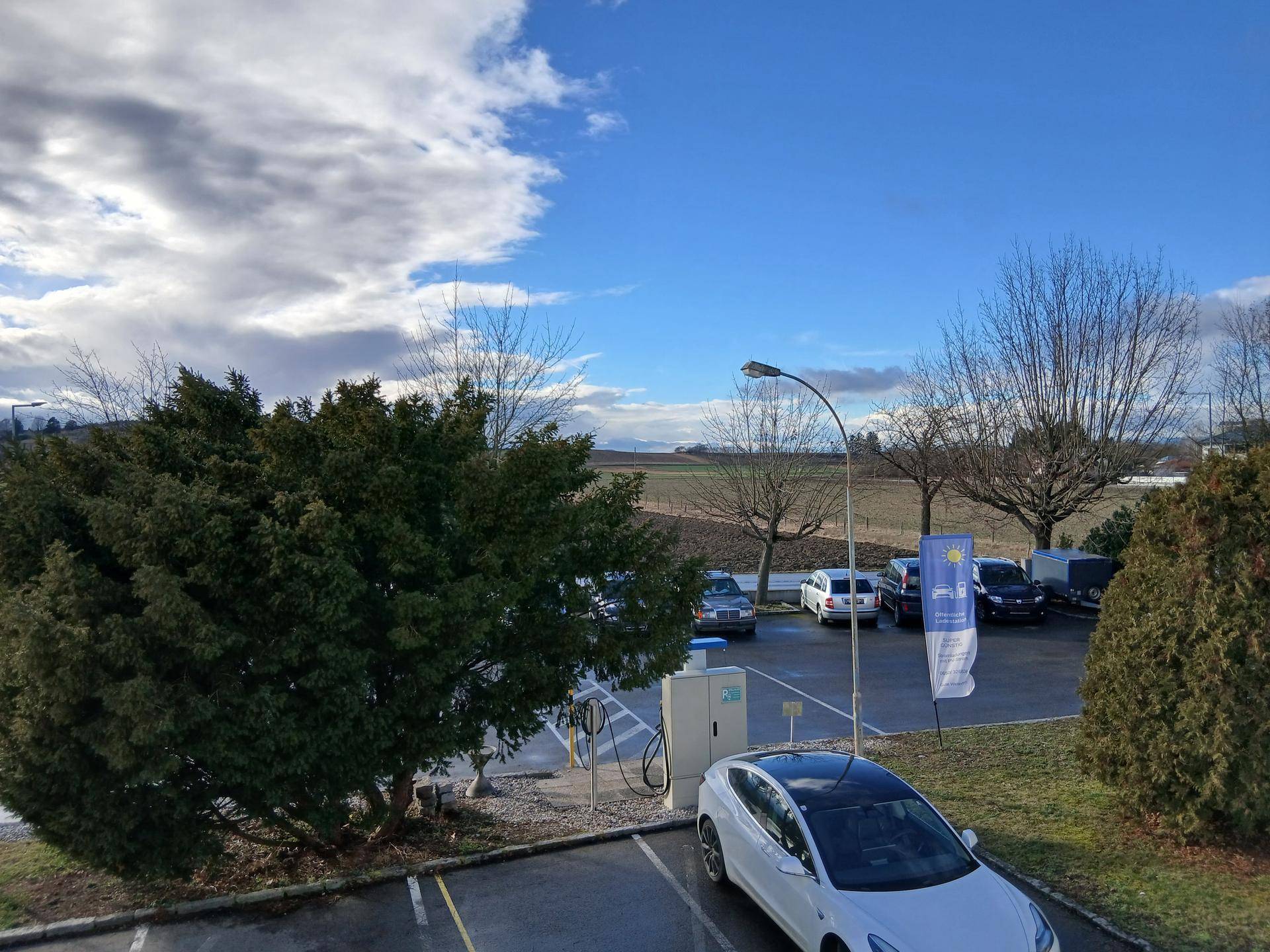 Außenparkplatz mit Ladestation für Elektrofahrzeuge, Bäumen und weitem Blick auf die Landschaft.