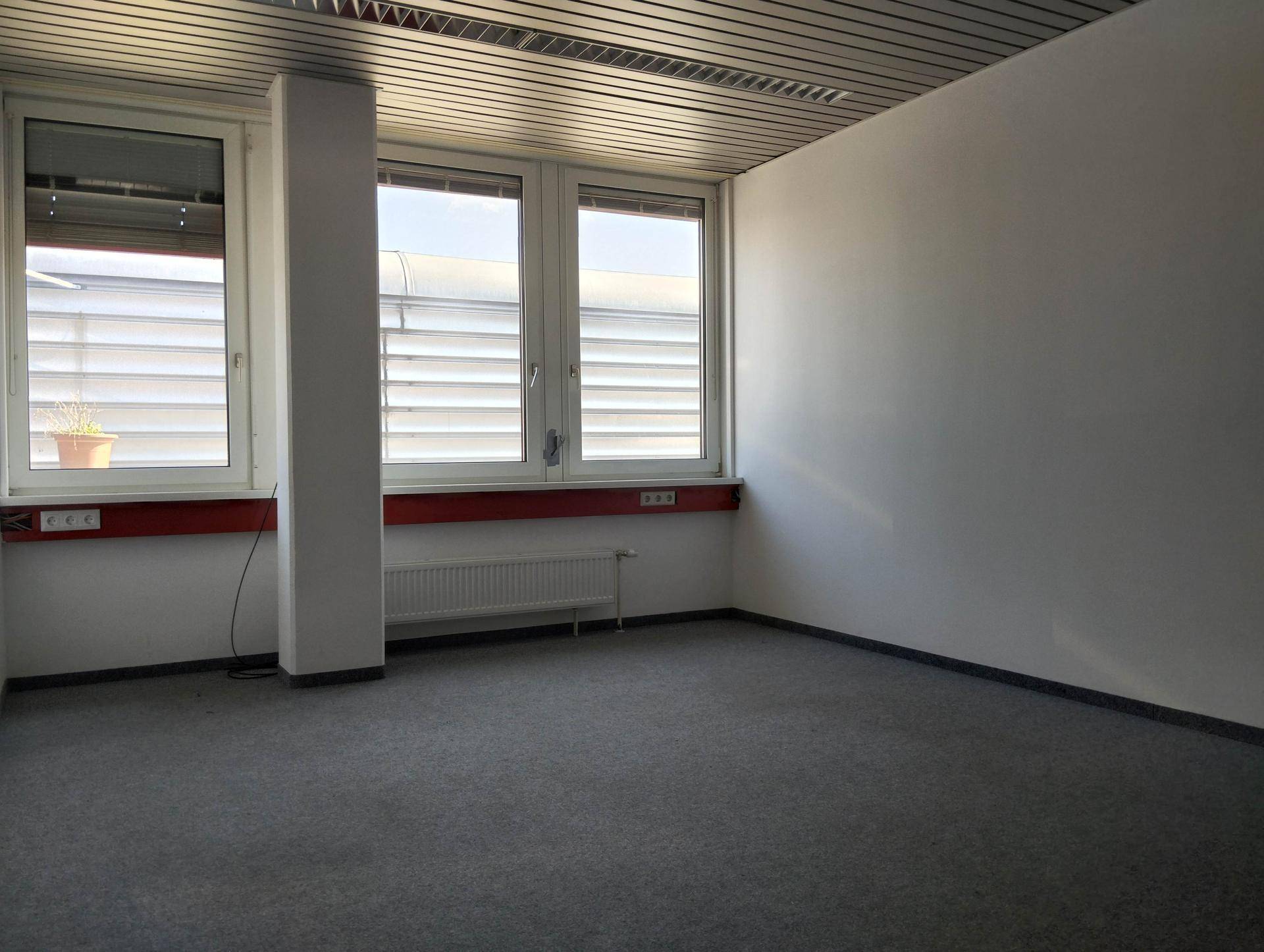 Leerer Büroraum mit grauem Teppichboden, weißen Wänden und Fenstern mit Jalousien, bereit zur individuellen Gestaltung.