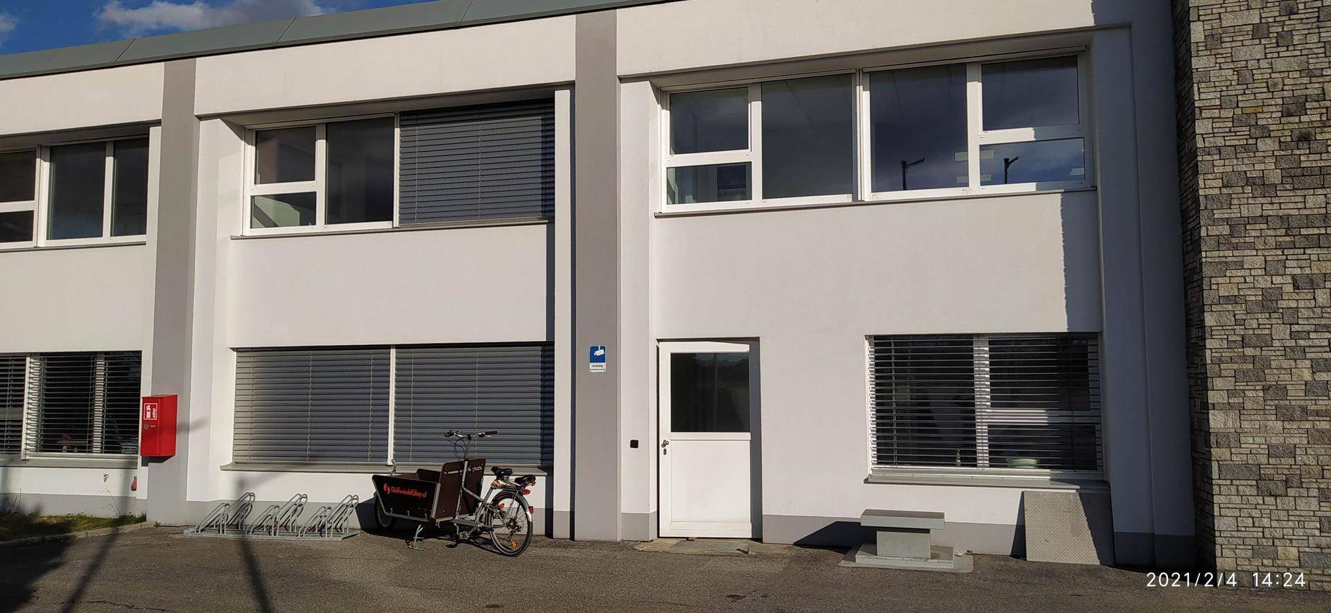 Moderne Gebäudefassade mit großen Fenstern, Jalousien und einem Fahrradständer davor.