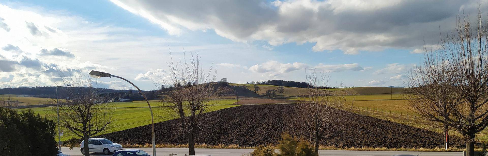 Panoramablick auf eine weite ländliche Landschaft mit Feldern, Hügeln und Bäumen unter bewölktem Himmel.