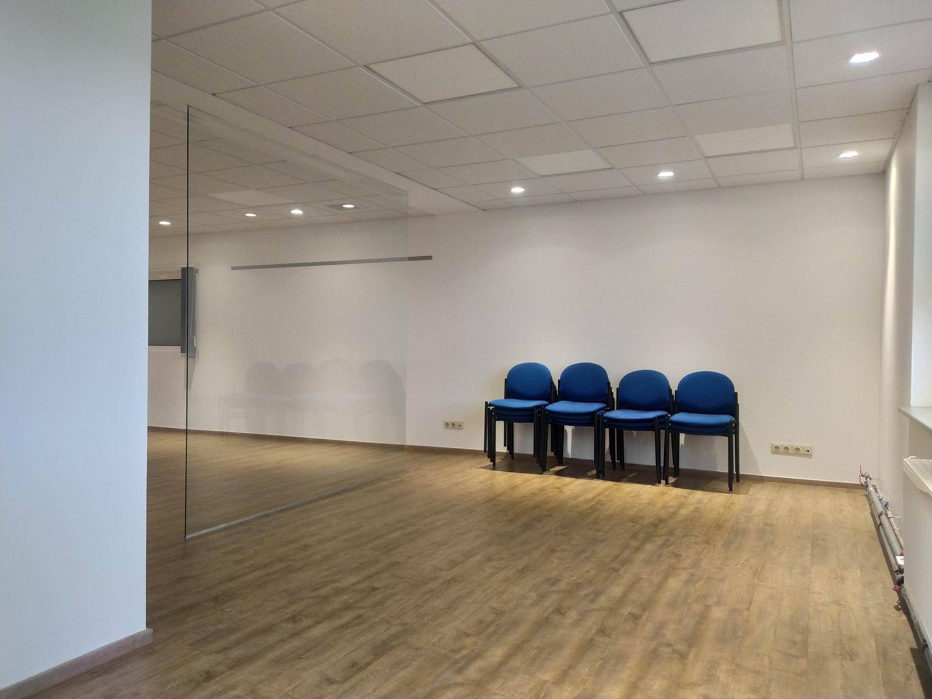 Heller Büroraum mit Laminatboden, weißen Wänden, blauen Stühlen und einer Glasabtrennung.