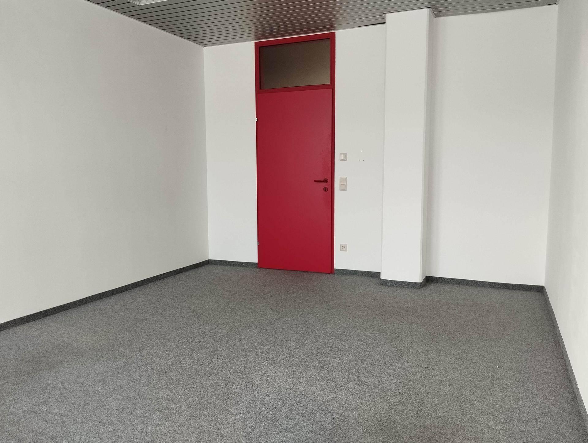 Heller, leerer Raum mit grauem Teppichboden und einer auffälligen roten Tür, ideal für Bürozwecke.