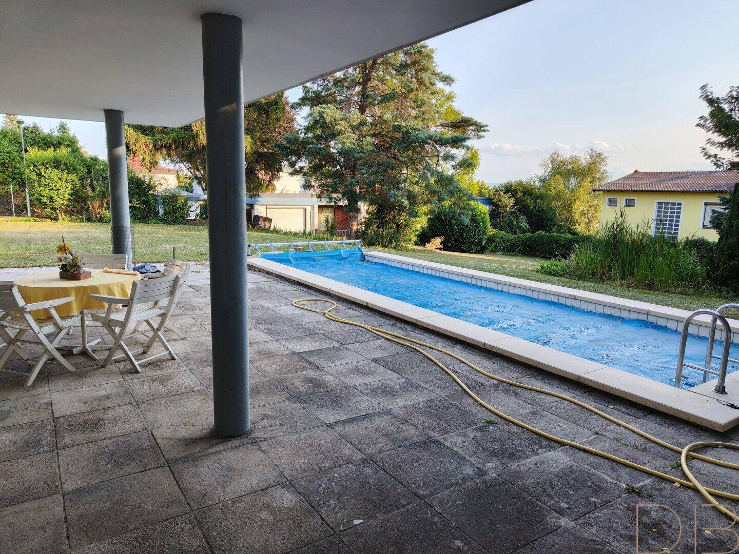 Große Terrasse mit Pool und Sitzgelegenheiten, umgeben von viel Grün.