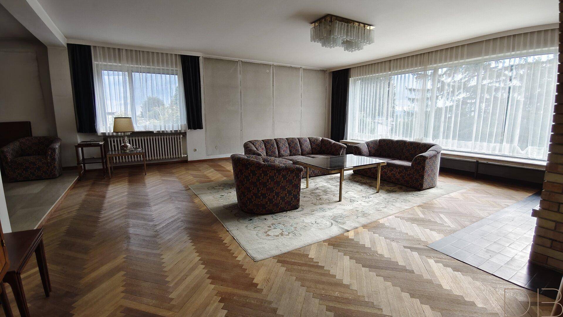 Geräumiges Wohnzimmer mit Fischgrätparkett, großen Fenstern und klassischer Einrichtung.