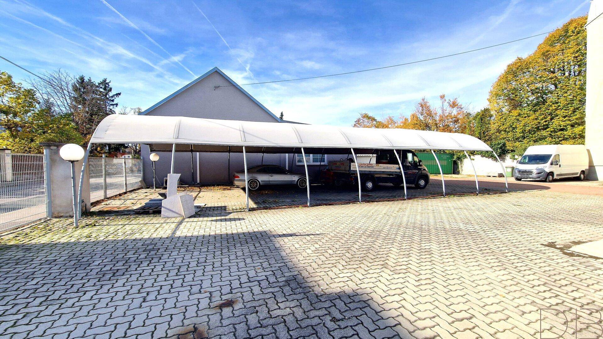 Außenansicht des Grundstücks mit einem langen Carport, der mehrere Fahrzeuge schützt, unter blauem Himmel.