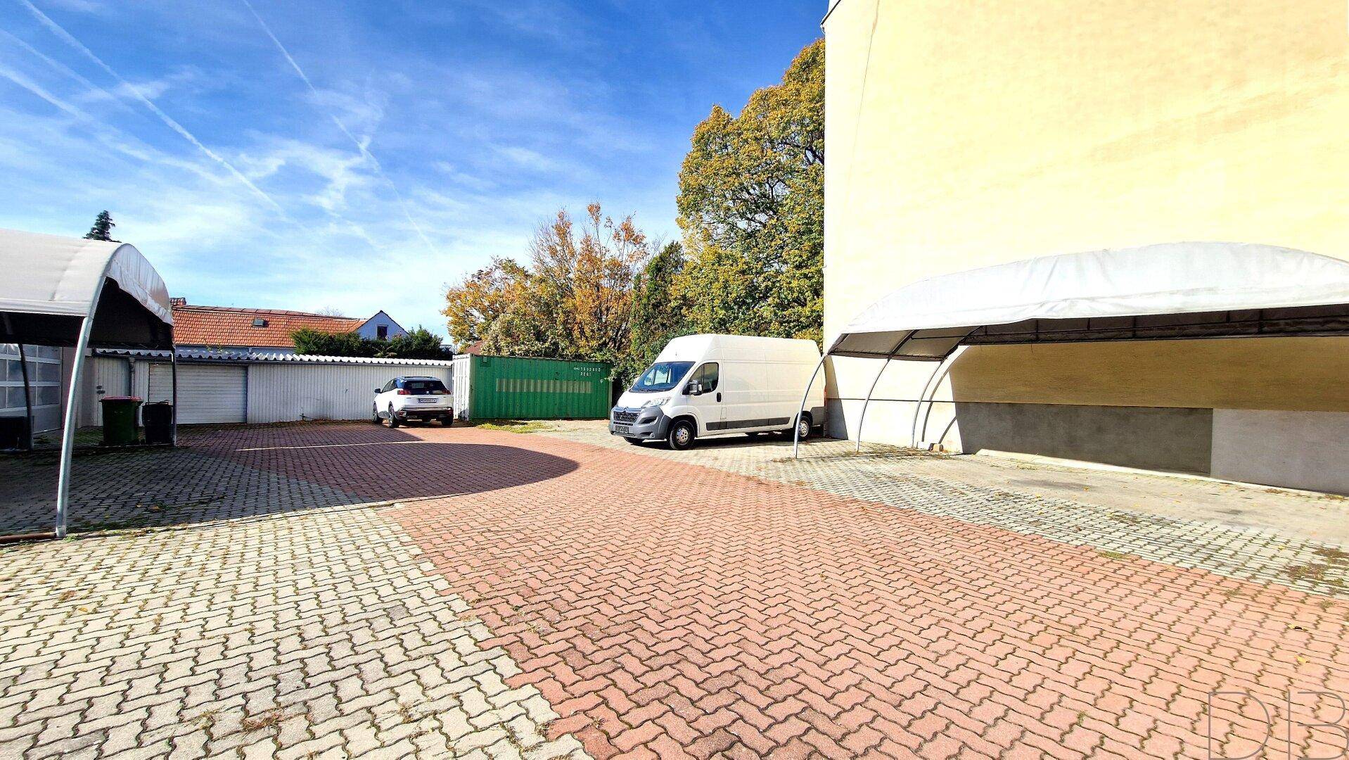Gepflasterter Außenbereich mit Parkplätzen, einem weißen Lieferwagen und einem weiteren Carport unter strahlendem Himmel.