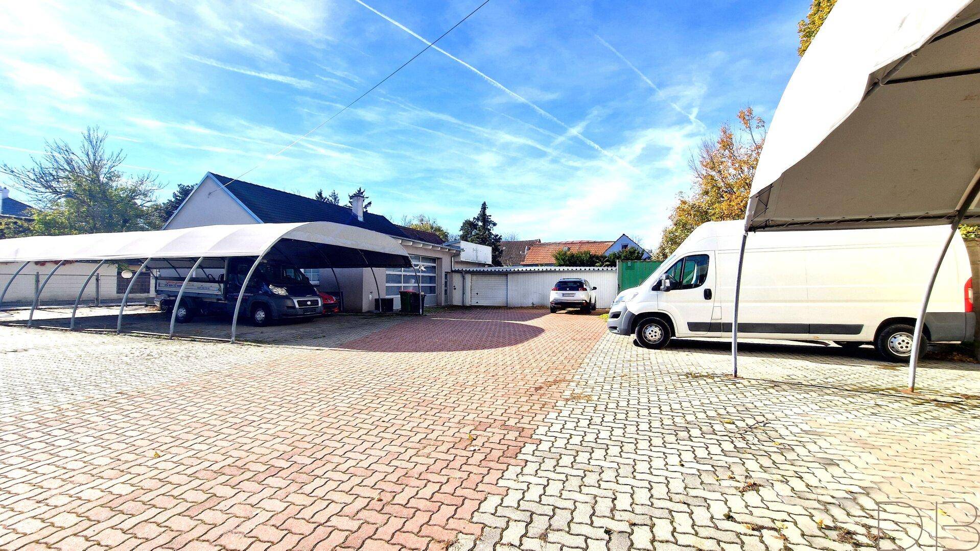 Weitläufiger Außenbereich mit gepflasterten Parkflächen, einem langen Carport und mehreren Fahrzeugen an einem sonnigen Tag.