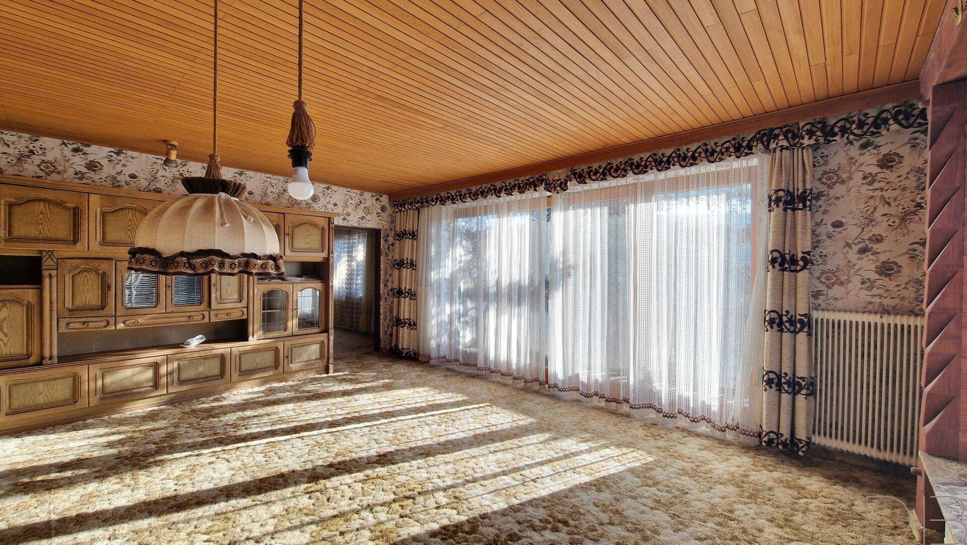 Helles Wohnzimmer mit Teppichboden, Holzdecke und floraler Tapete, durchflutet von Sonnenlicht.