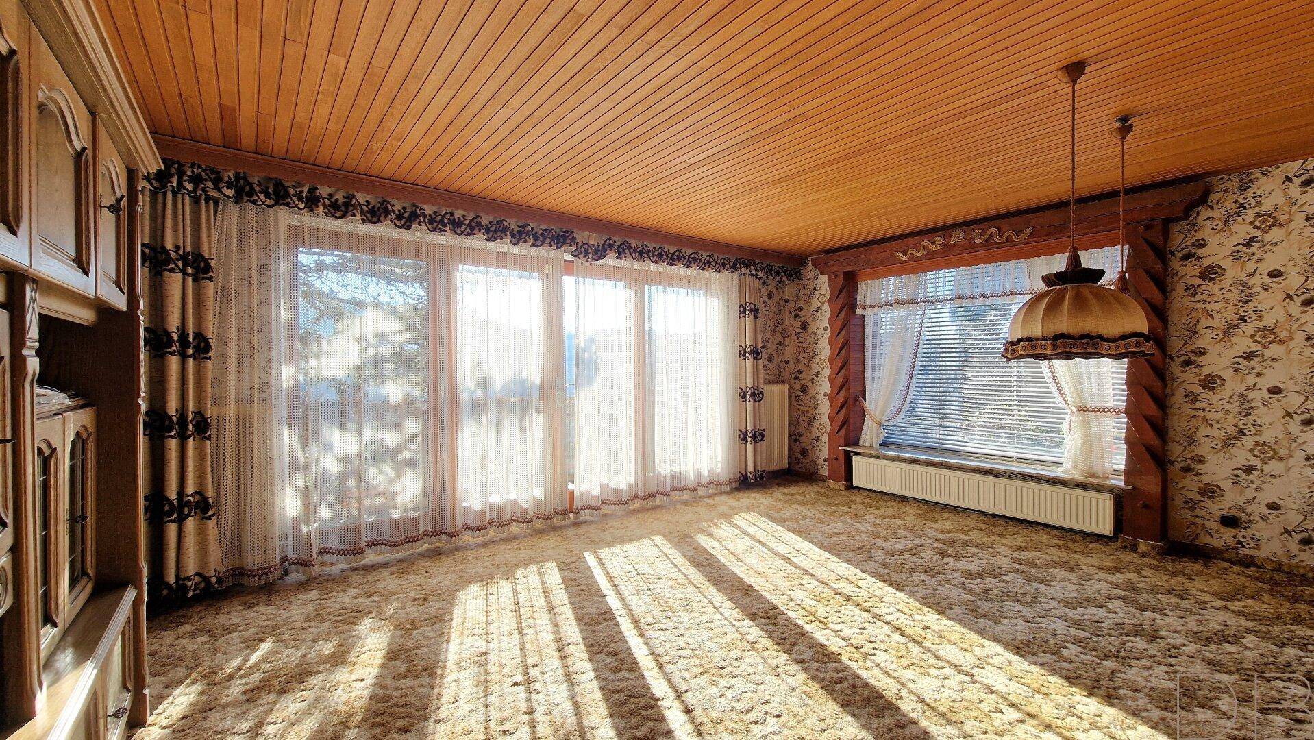 Geräumiges Wohnzimmer mit Teppichboden, Holzdecke und großen Fenstern, die viel Licht hereinlassen.