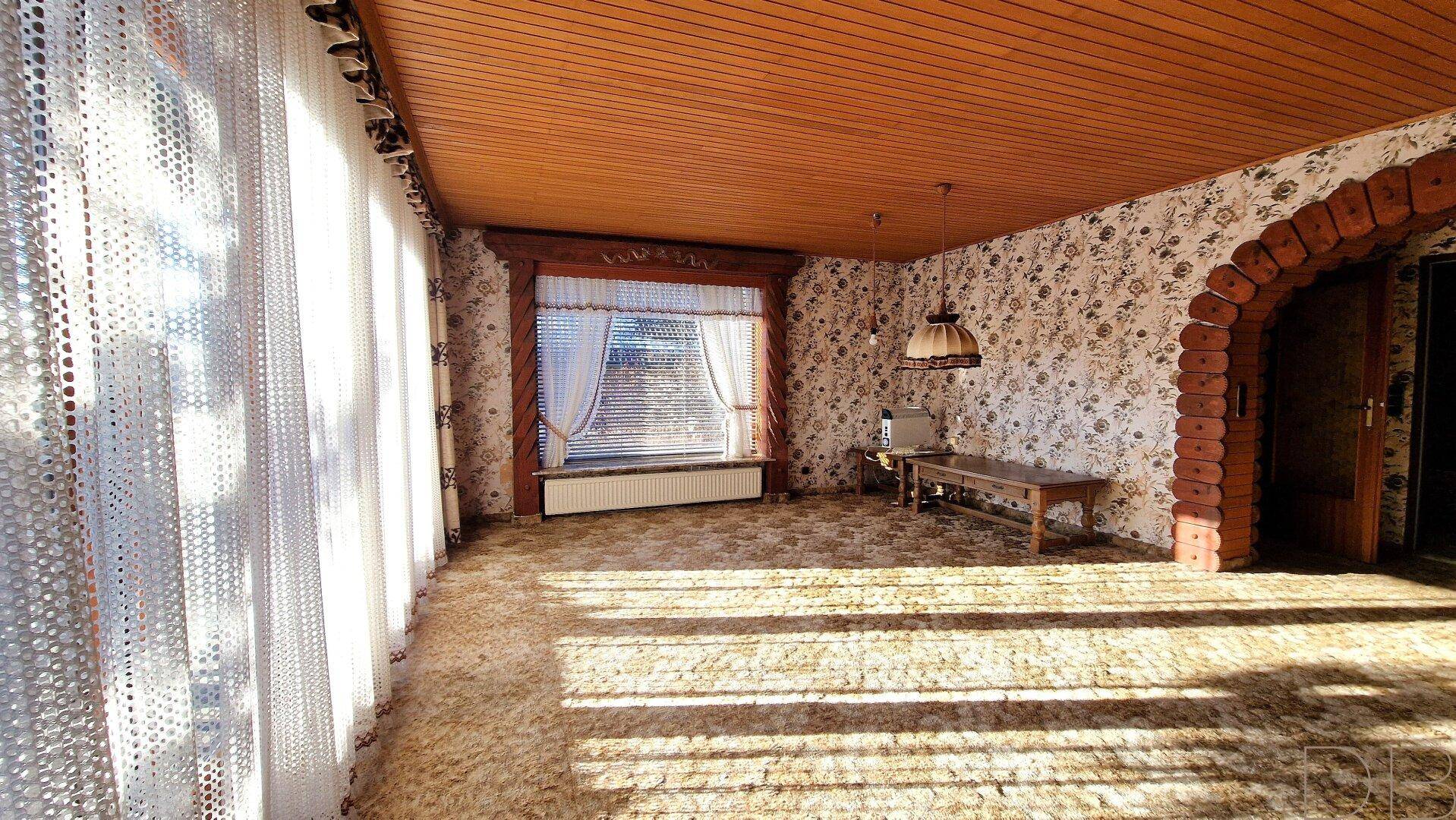 Geräumiges Wohnzimmer mit Teppichboden, Holzdecke und einem dekorativen Rundbogen.