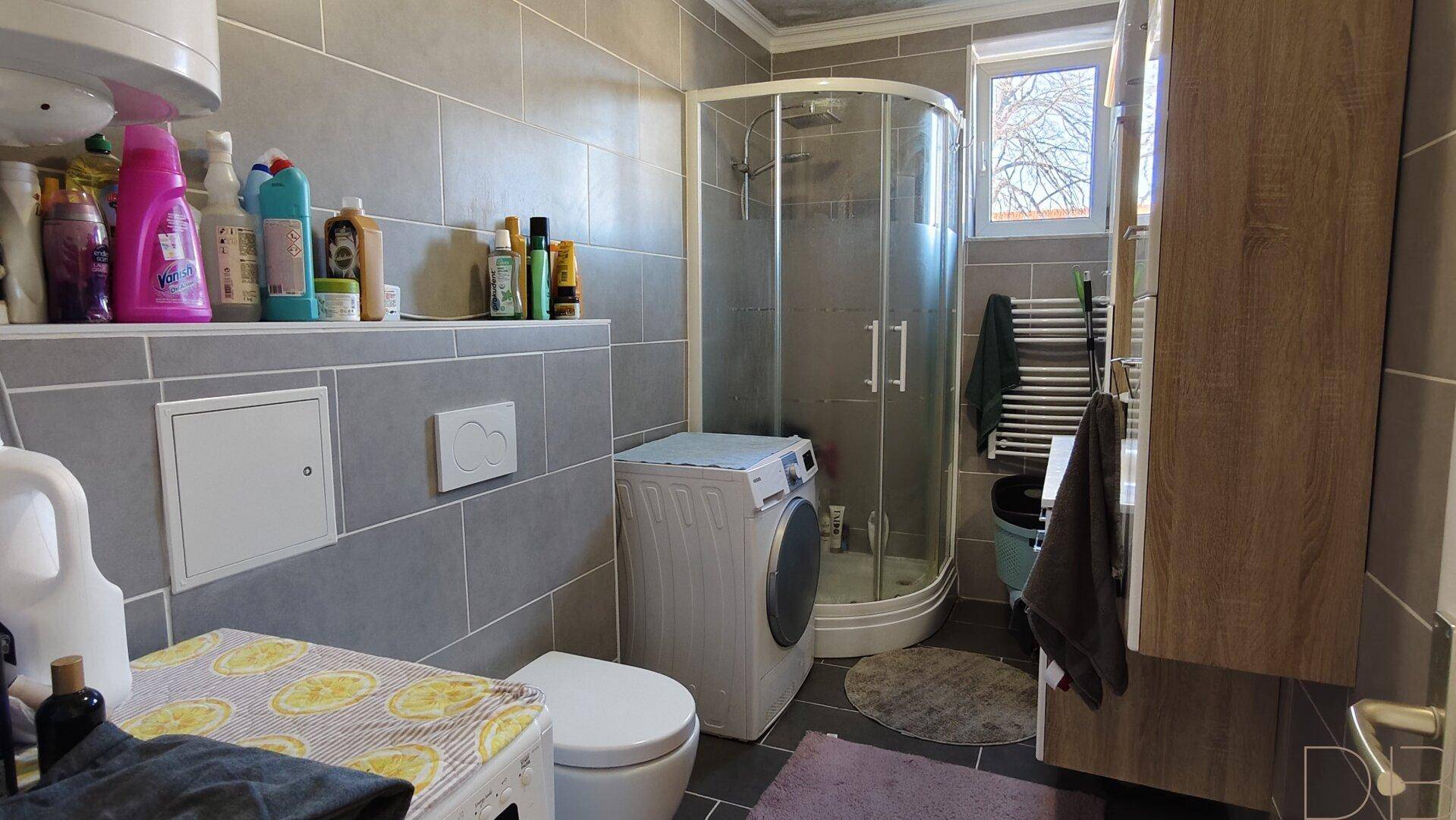 Modernes Badezimmer mit Duschkabine, Waschmaschine und grauen Fliesen, praktisch eingerichtet.