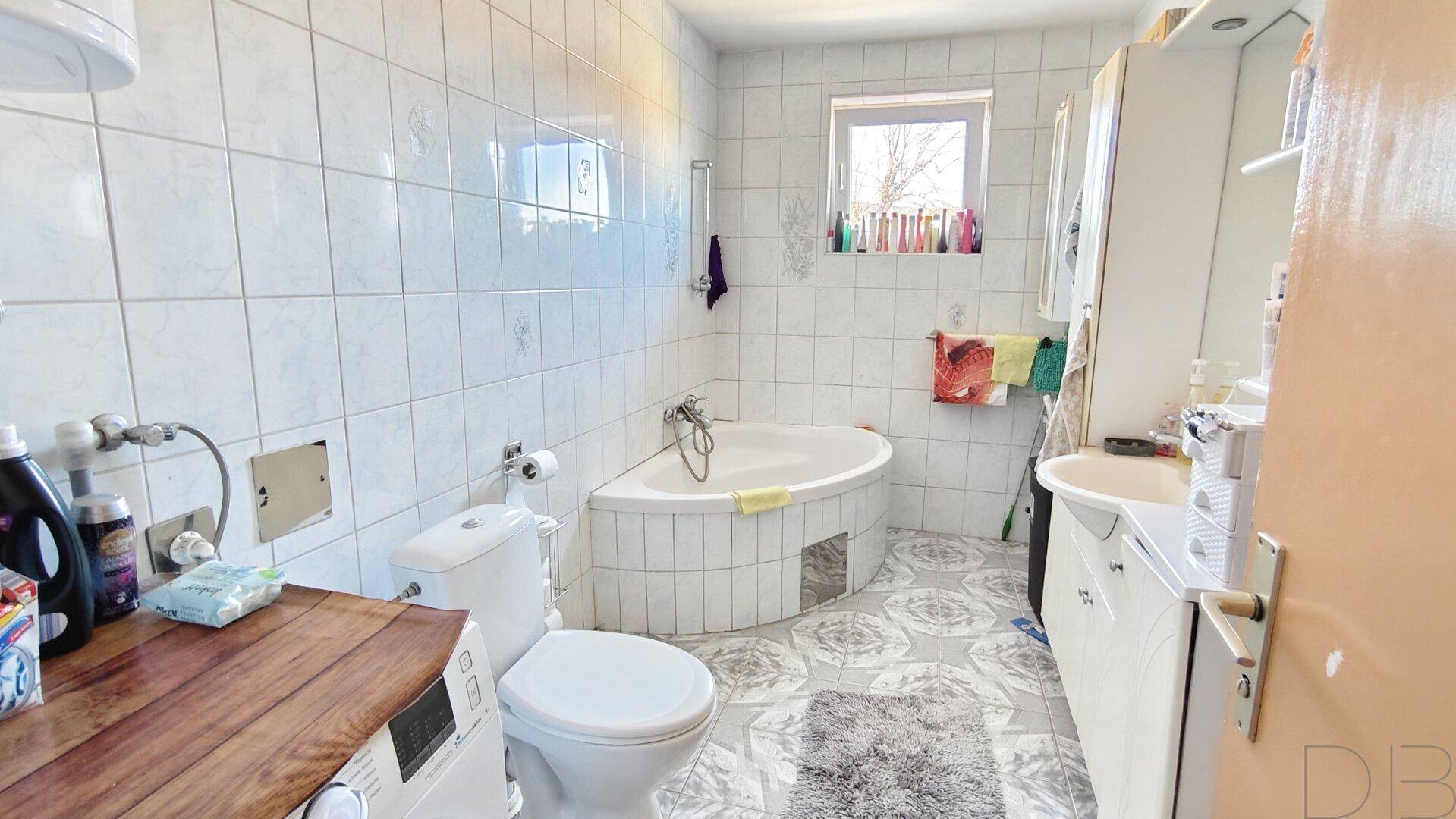 Badezimmer mit Eckbadewanne, weißen Fliesen und Fenster, hell und funktional.