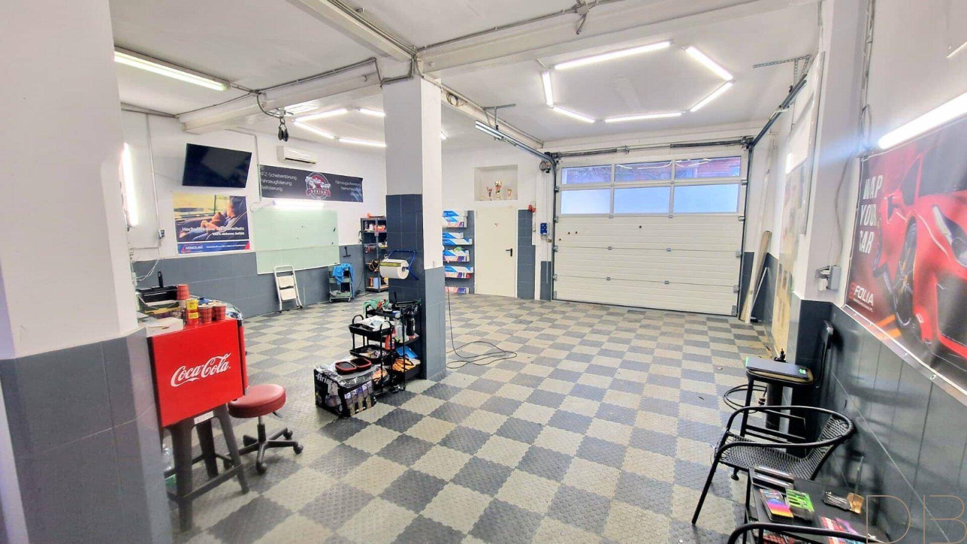 Innenansicht einer geräumigen Garage oder Werkstatt mit großem Rolltor, kariertem Boden und Arbeitsbereichen.