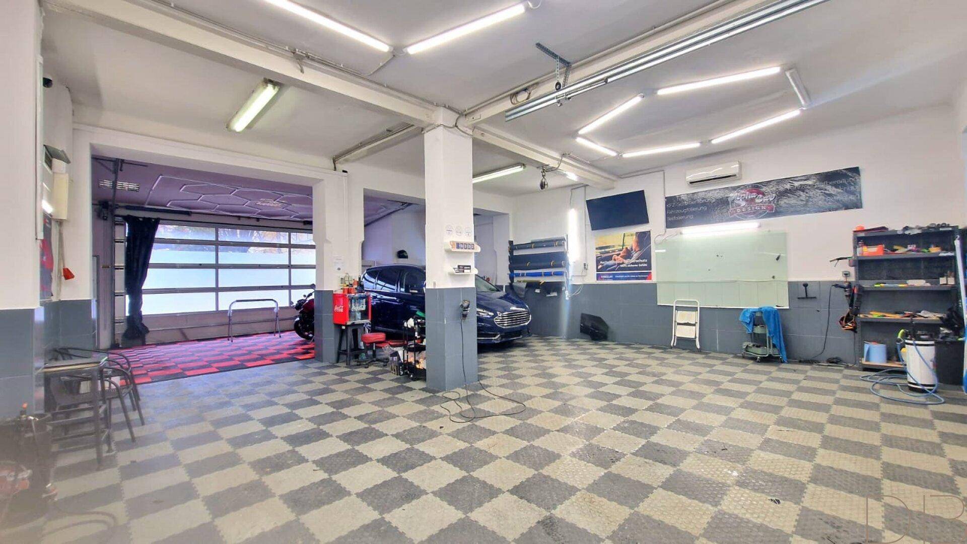 Geräumige Werkstatt oder Garage mit kariertem Boden, Rolltor und heller Deckenbeleuchtung.