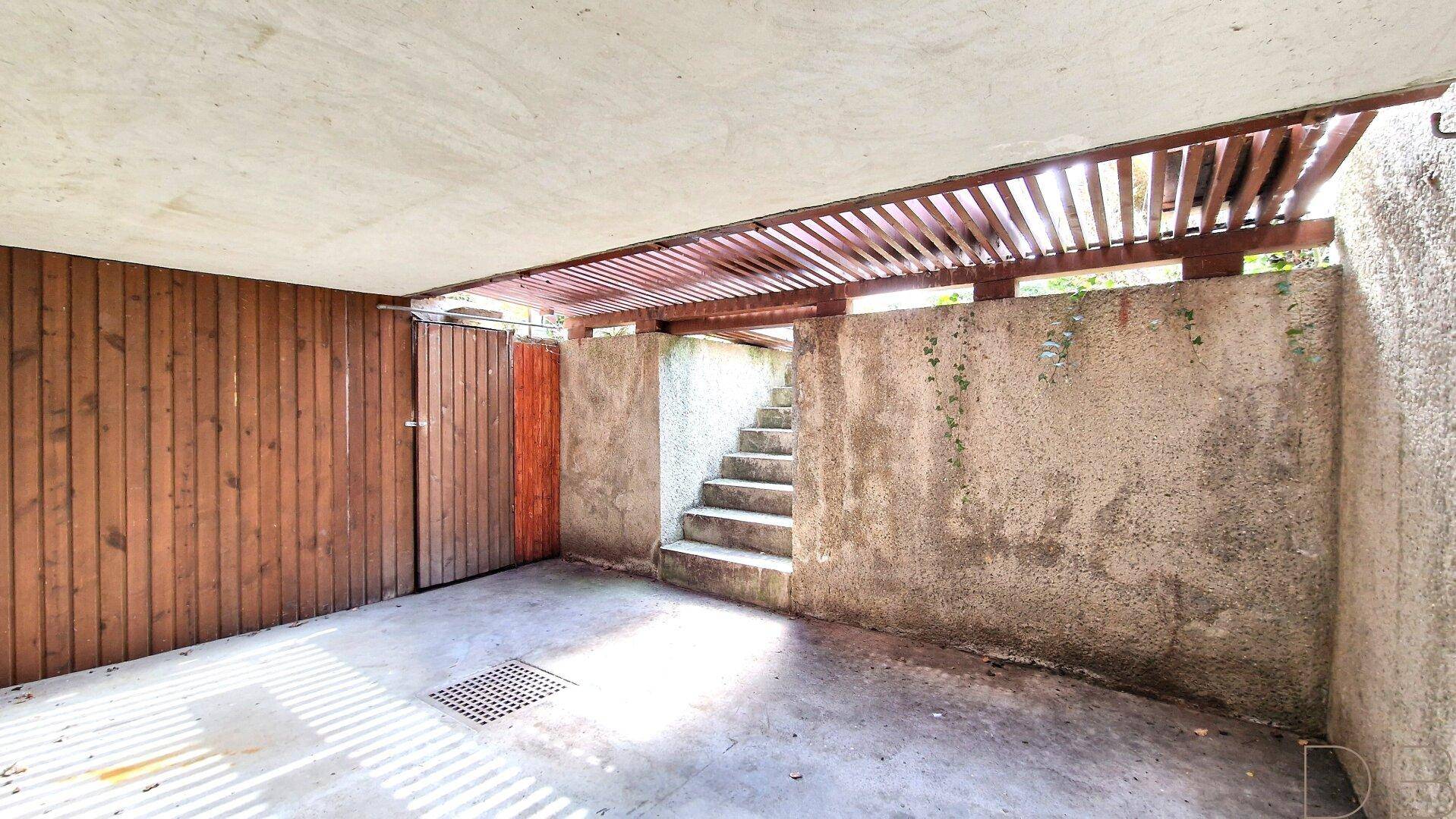 Überdachter Garagenbereich mit Holztor und Betonwänden, bietet Schutz für Fahrzeuge.
