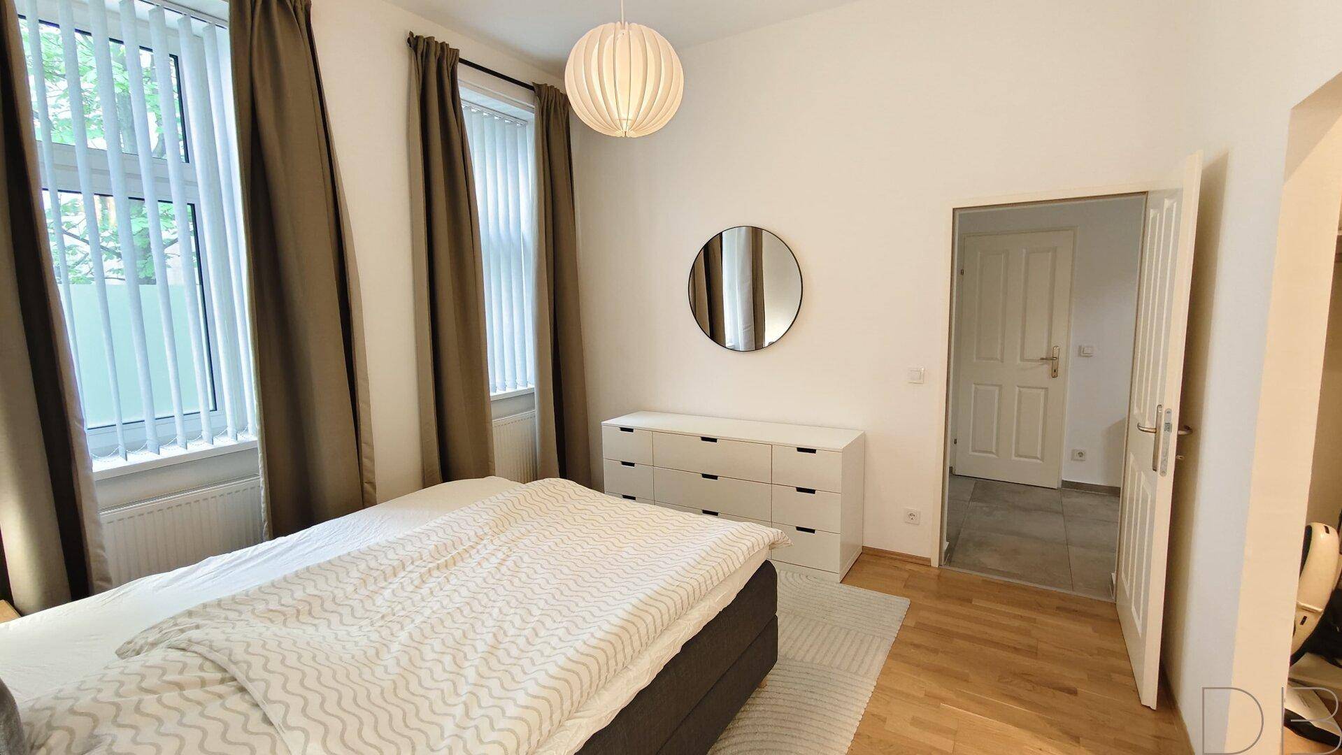 Helles Schlafzimmer mit Doppelbett, Kommode und großem Spiegel.