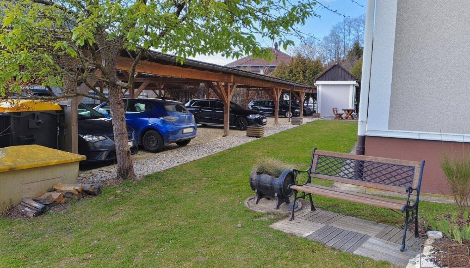 Gepflegter Gartenbereich mit Sitzbank, Bäumen und einem überdachten Carport für mehrere Fahrzeuge.