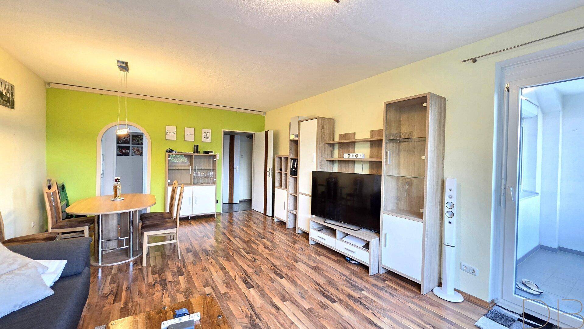 Geräumiger Wohn-Essbereich mit Holzboden, grüner Akzentwand und Zugang zum Balkon.