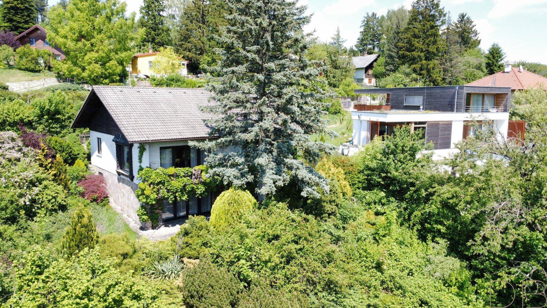 Einfamilienhaus mit überdachter Loggia, umgeben von üppigem Grün und einem modernen Nachbargebäude.