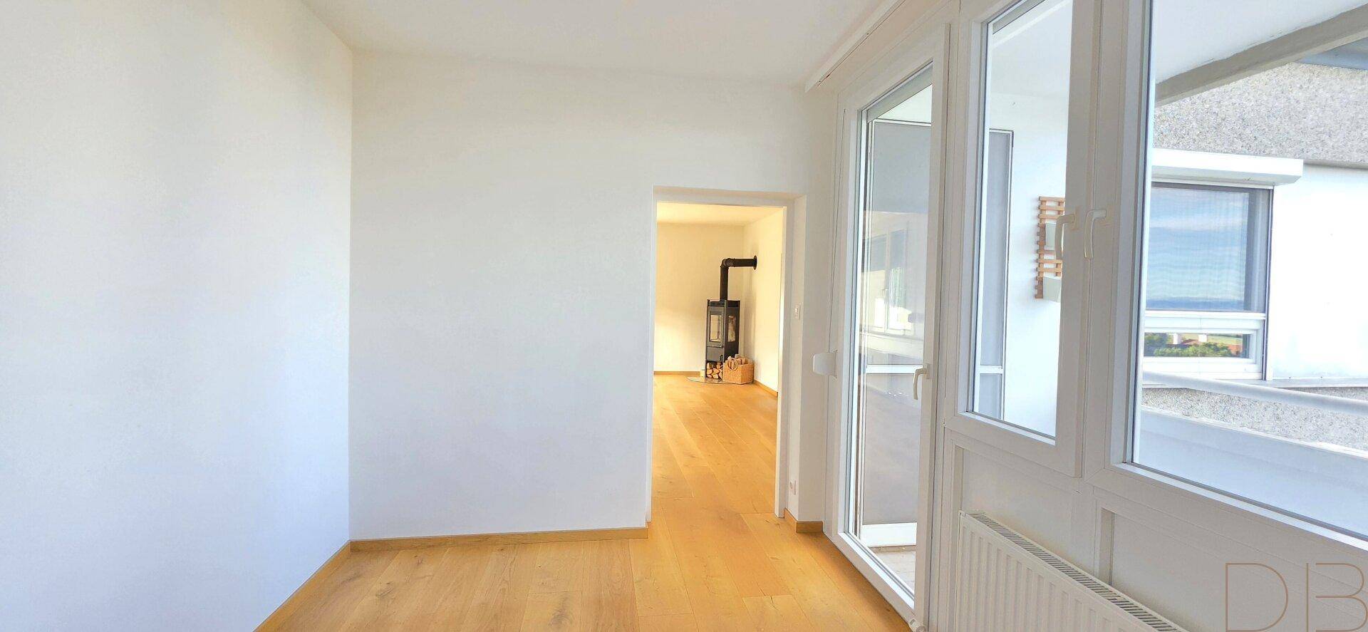 Heller Raum mit Holzboden und Zugang zum Balkon, Kaminofen im Hintergrund sichtbar.