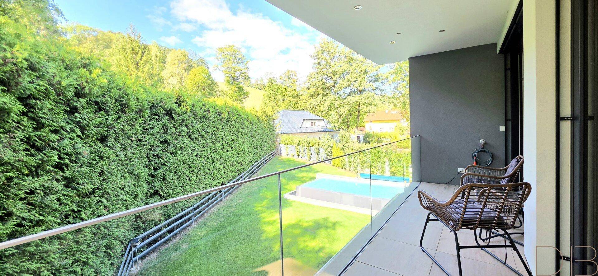 Langer Balkon mit Glasgeländer, Sitzgelegenheiten und Blick auf den Pool und die Natur.