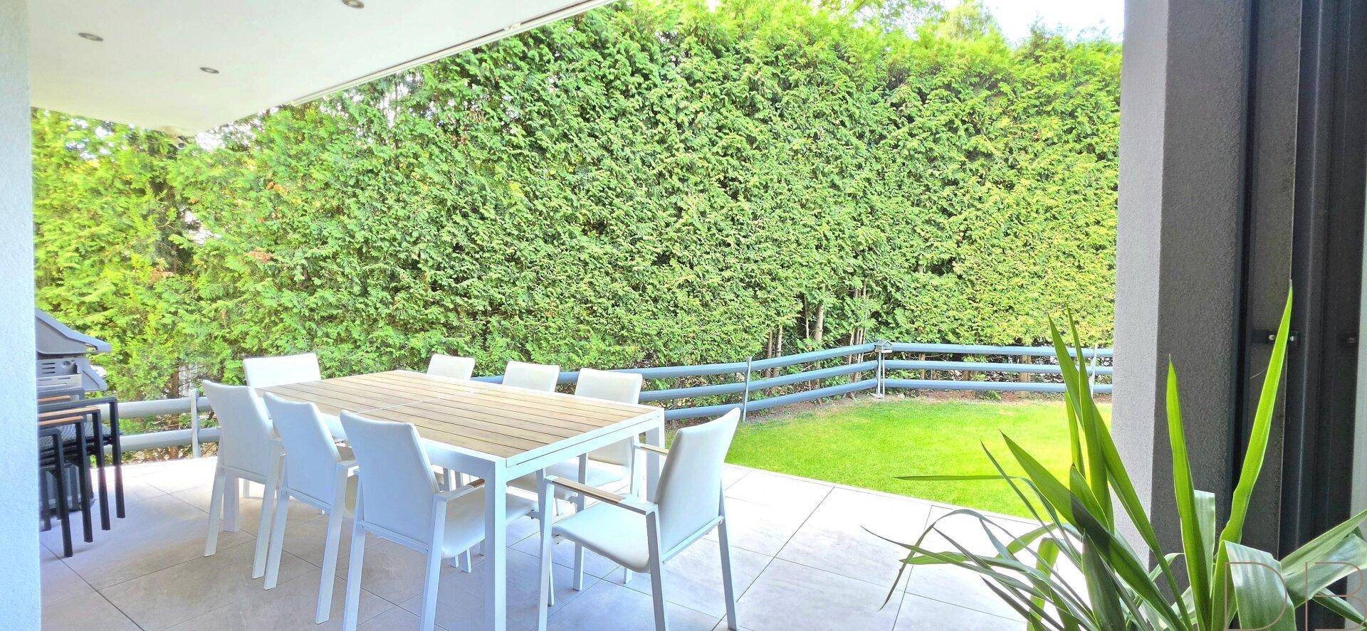 Große überdachte Terrasse mit Esstisch und Stühlen, umgeben von einer hohen Hecke.
