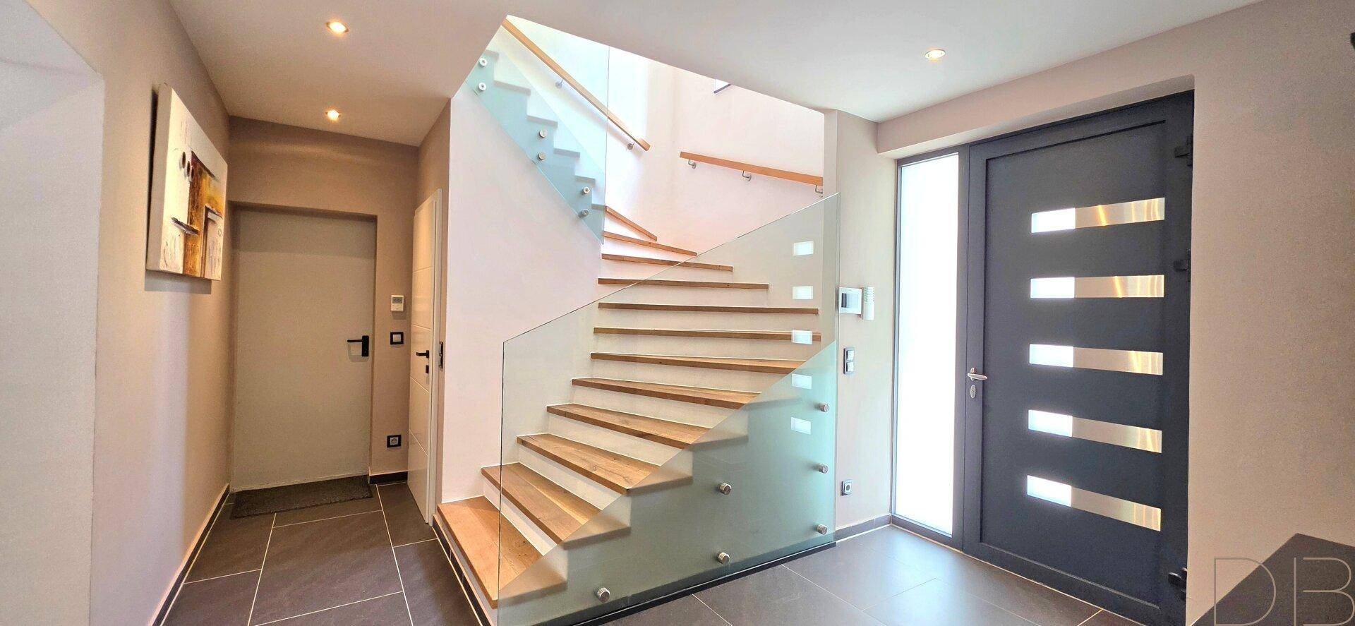 Moderner Eingangsbereich mit eleganter Holztreppe, Glasgeländer und dunkler Eingangstür.