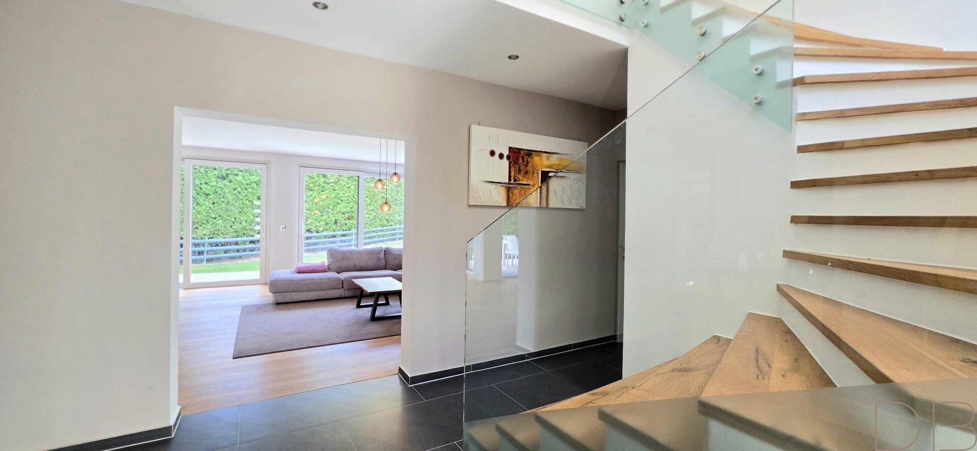Heller Eingangsbereich mit moderner Holztreppe, Glasgeländer und Blick ins Wohnzimmer.
