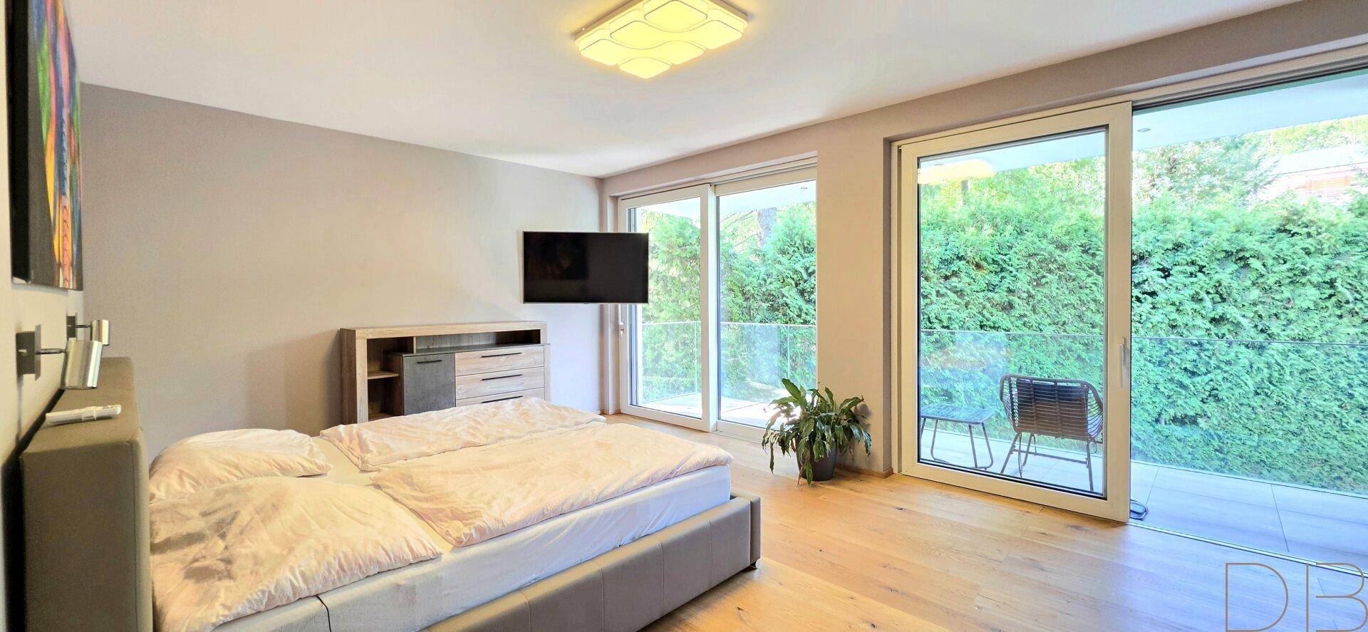 Schlafzimmer mit Holzboden, großem Bett und Zugang zu einem Balkon mit Heckenblick.