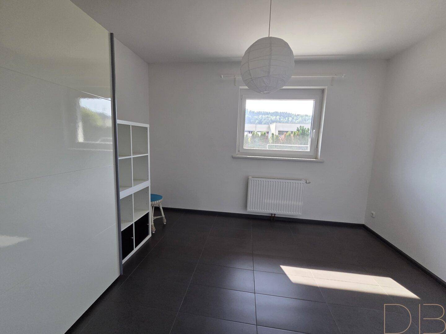 Helles Schlafzimmer mit dunklen Fliesen, weißem Schrank und Fenster mit Blick ins Grüne.