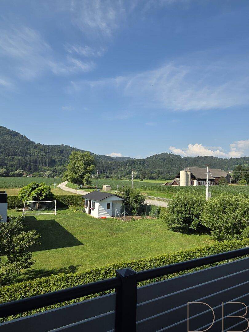 Balkon mit weitem Blick über grüne Felder und bewaldete Hügel unter blauem Himmel.